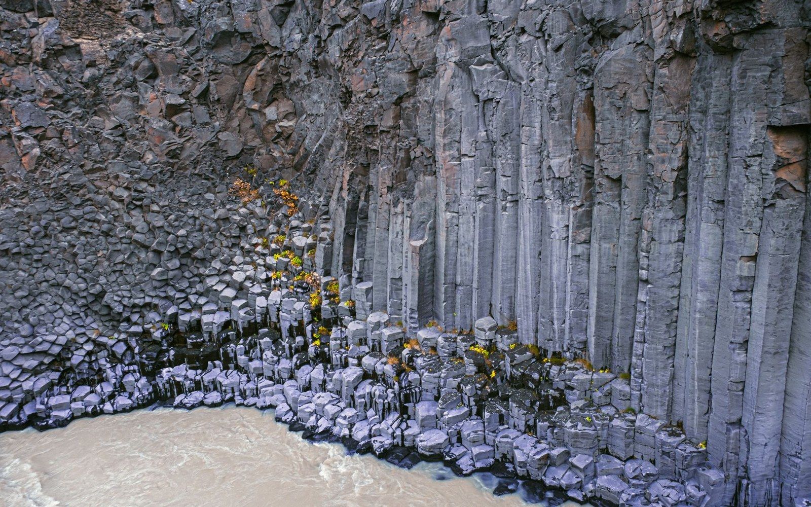 Hexagonal basalt columns rise above a muddy river, with small shrubs growing between the rocks
