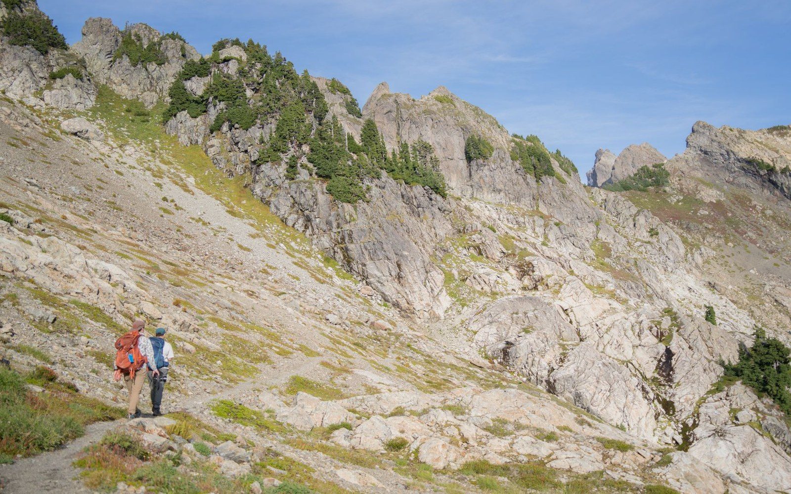 Hiker with a backpack walks a rocky trail below jagged, tree-dotted mountain ridges