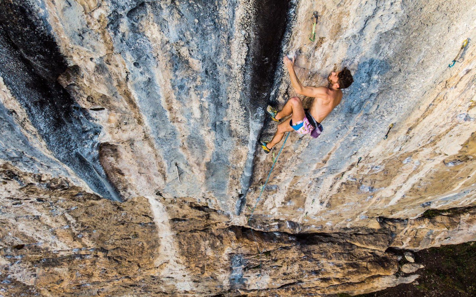 A shirtless climber ascends a steep limestone cliff using clipped rope protection