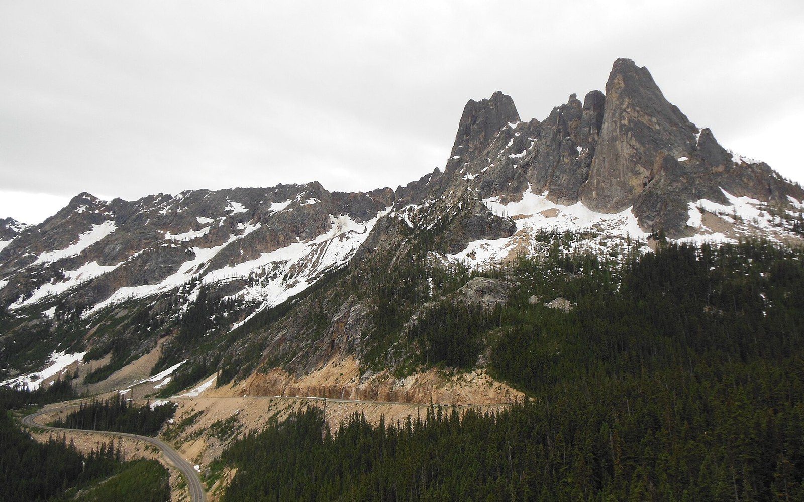 Jagged rocky peaks with patches of snow rise above a forested mountain highway