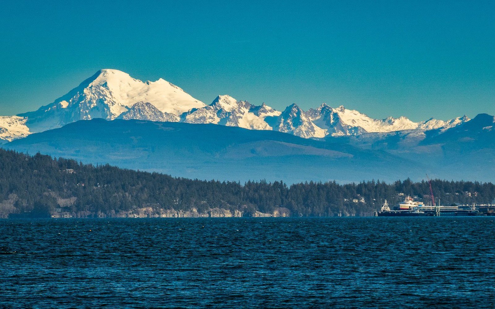 Snow-capped mountain range rising above forested coastline with a ferry crossing the strait