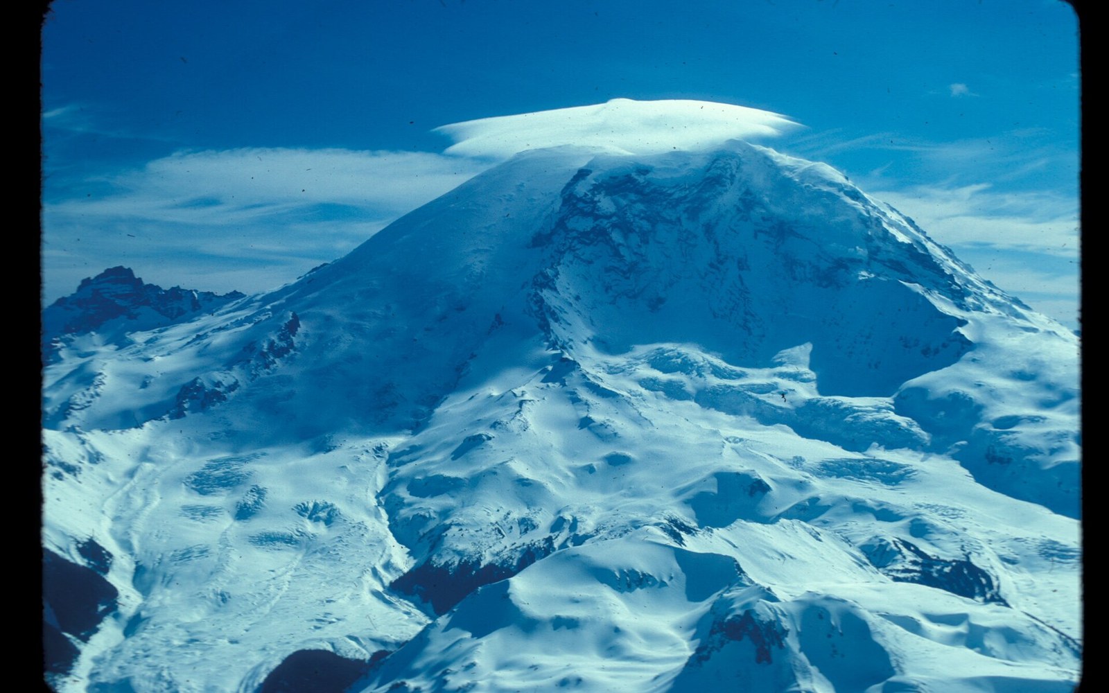 A lenticular cloud caps Mount Rainier's snow-covered summit above glaciated ridges
