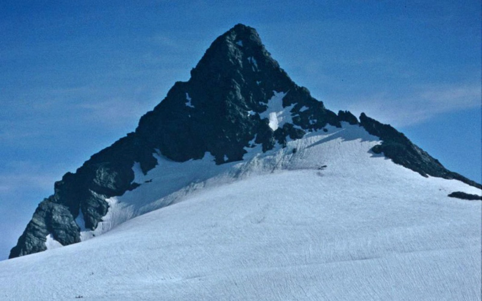 Snow-covered slopes lead up to a jagged, dark rocky summit under clear blue sky