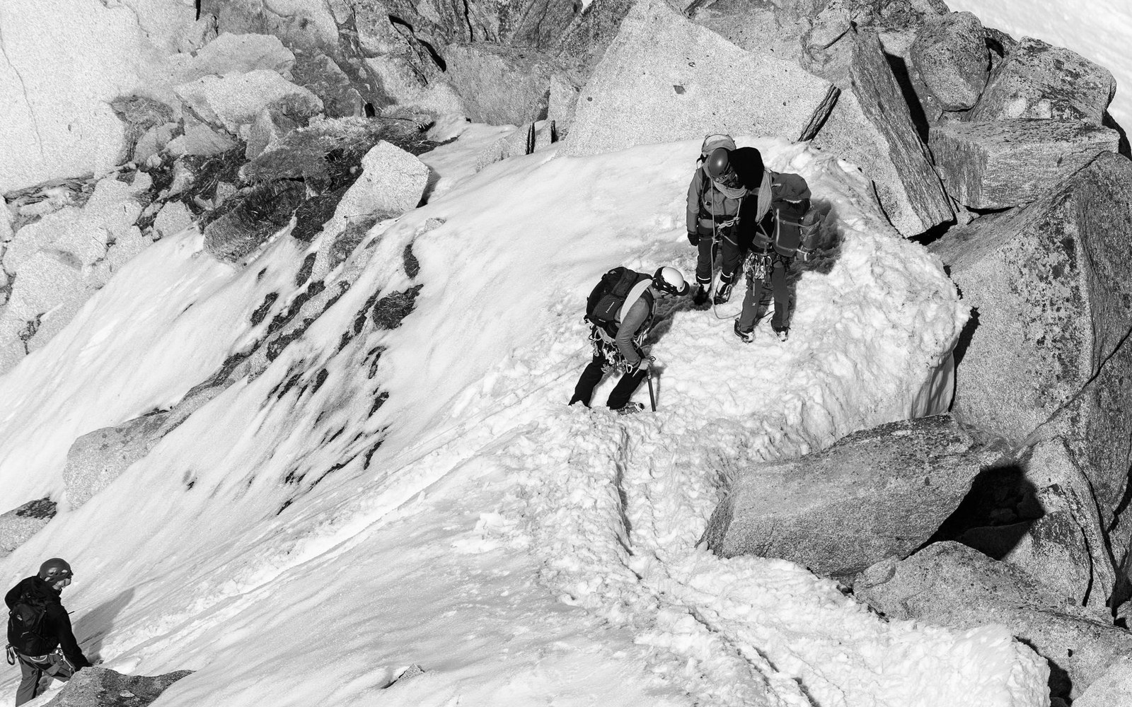 Climbers with ice axes and backpacks navigate a rocky, snow-covered ridge
