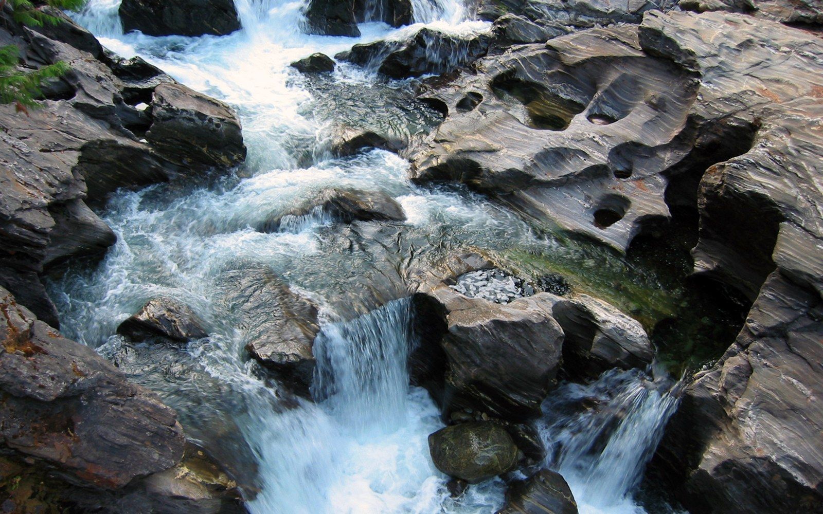 Rushing water carves smooth potholes into dark rock along a cascading mountain stream